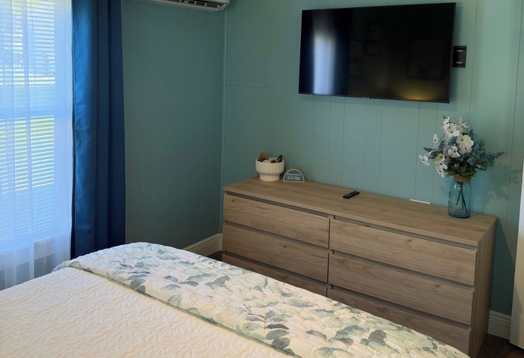 Ein Schlafzimmer mit einem Bett, einem Schminktisch, einem flachbildfernseher an der Wand und einem Fenster mit Vorhängen.