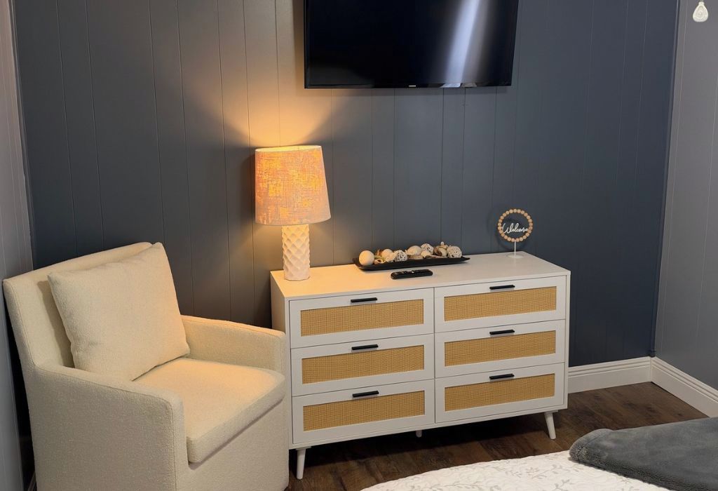Una habitación con un sillón blanco, un aparador blanco con cestos de mimbre, una lámpara y un televisor de pantalla plana montado en la pared.