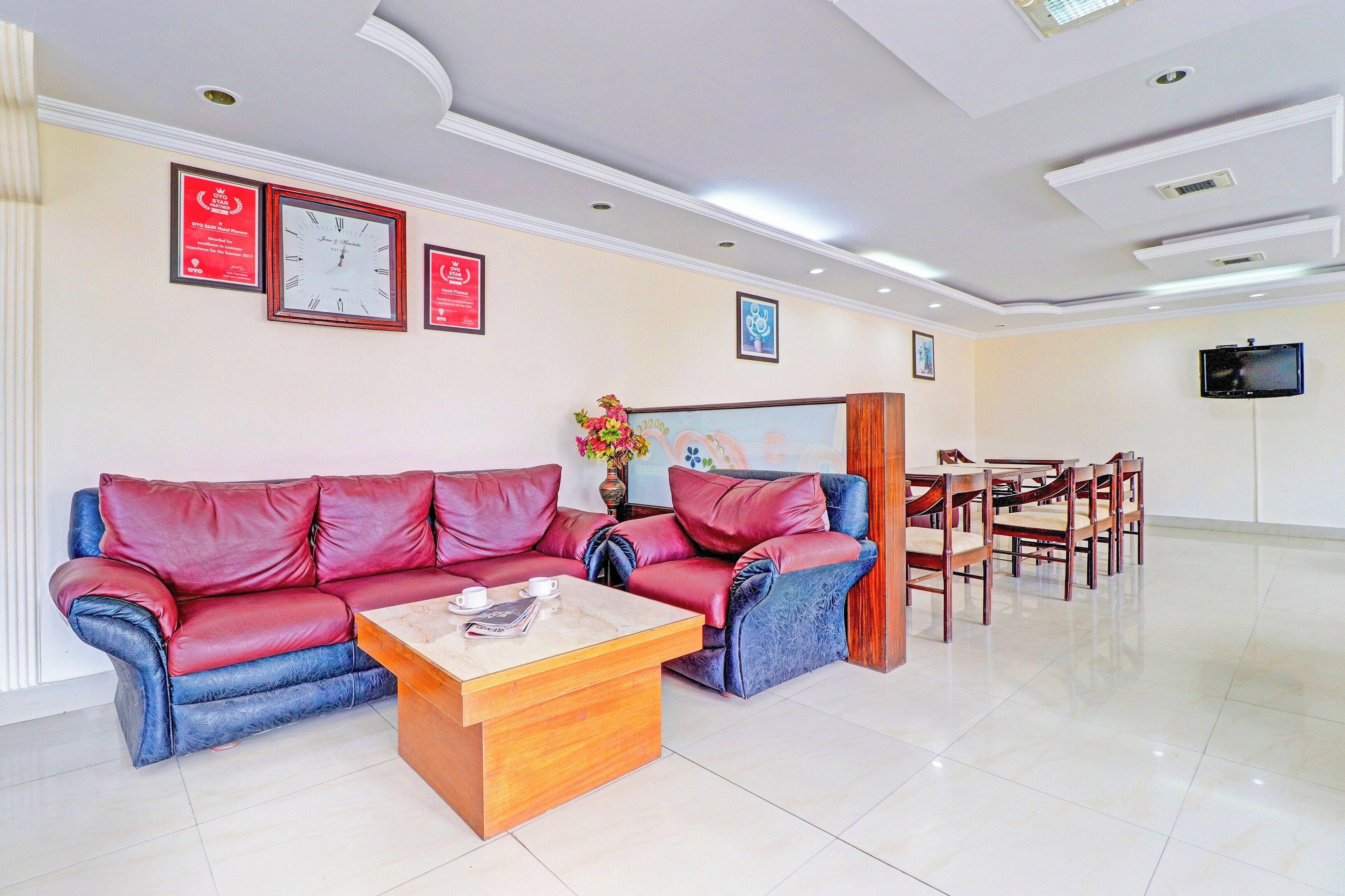 A hotel lobby with a red leather sofa, wooden coffee table, framed awards on the wall, and a television mounted on the wall.