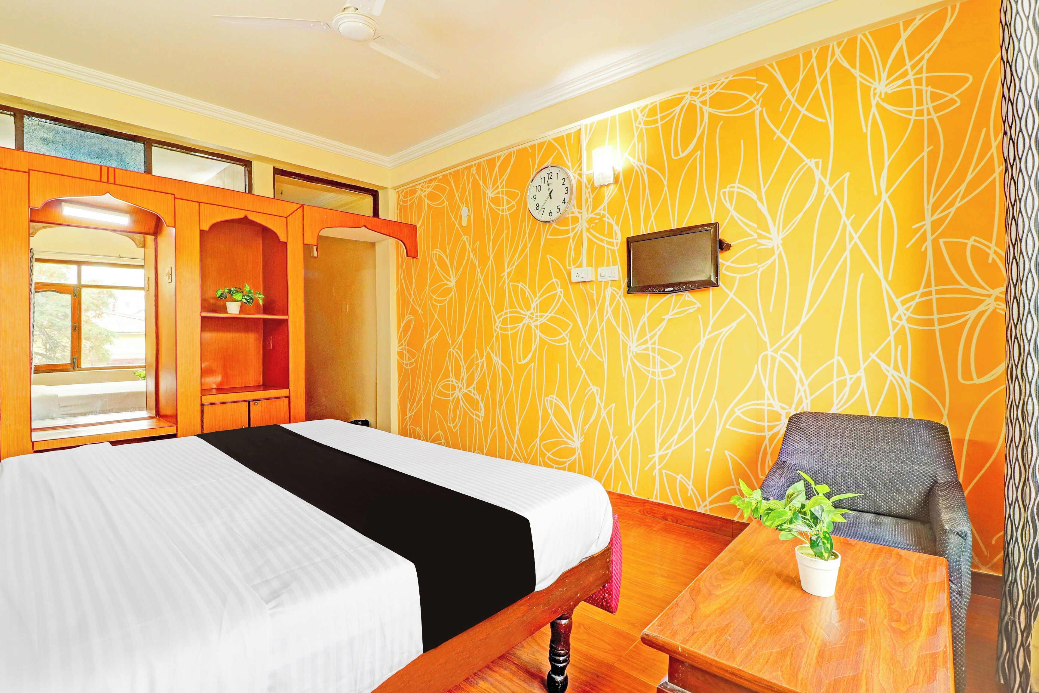 A hotel room with a bed, a chair, a small table, a TV, and a wall clock.