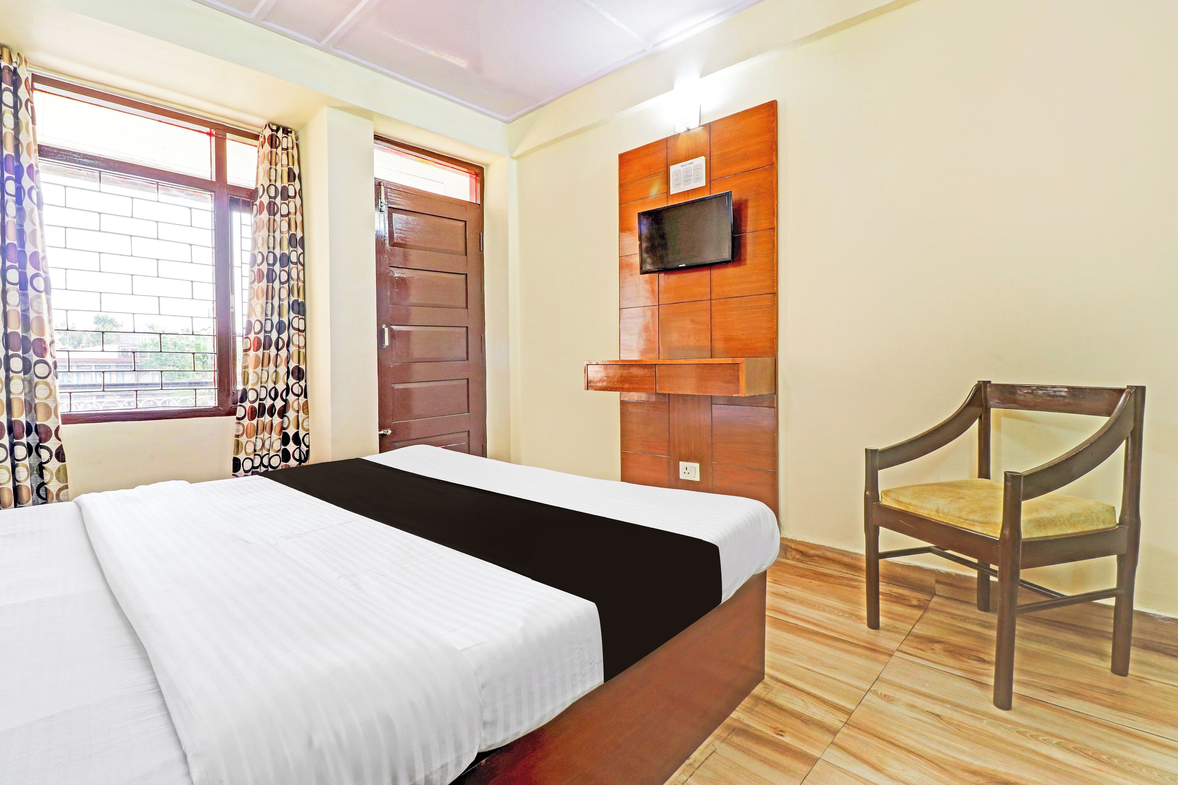 A hotel room with a bed, a chair, a television mounted on the wall, and a window with curtains.