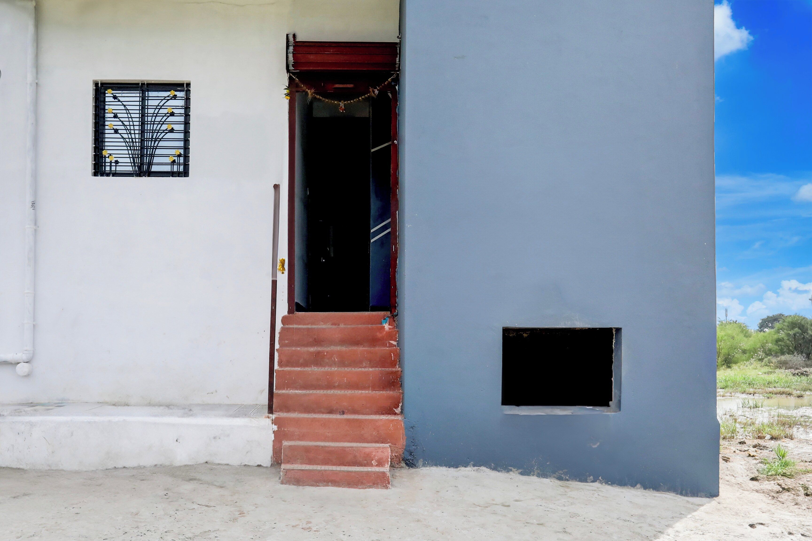 A building with a red staircase leading to a dark doorway, a small window with a metal grille, and a blue wall with a small square opening.