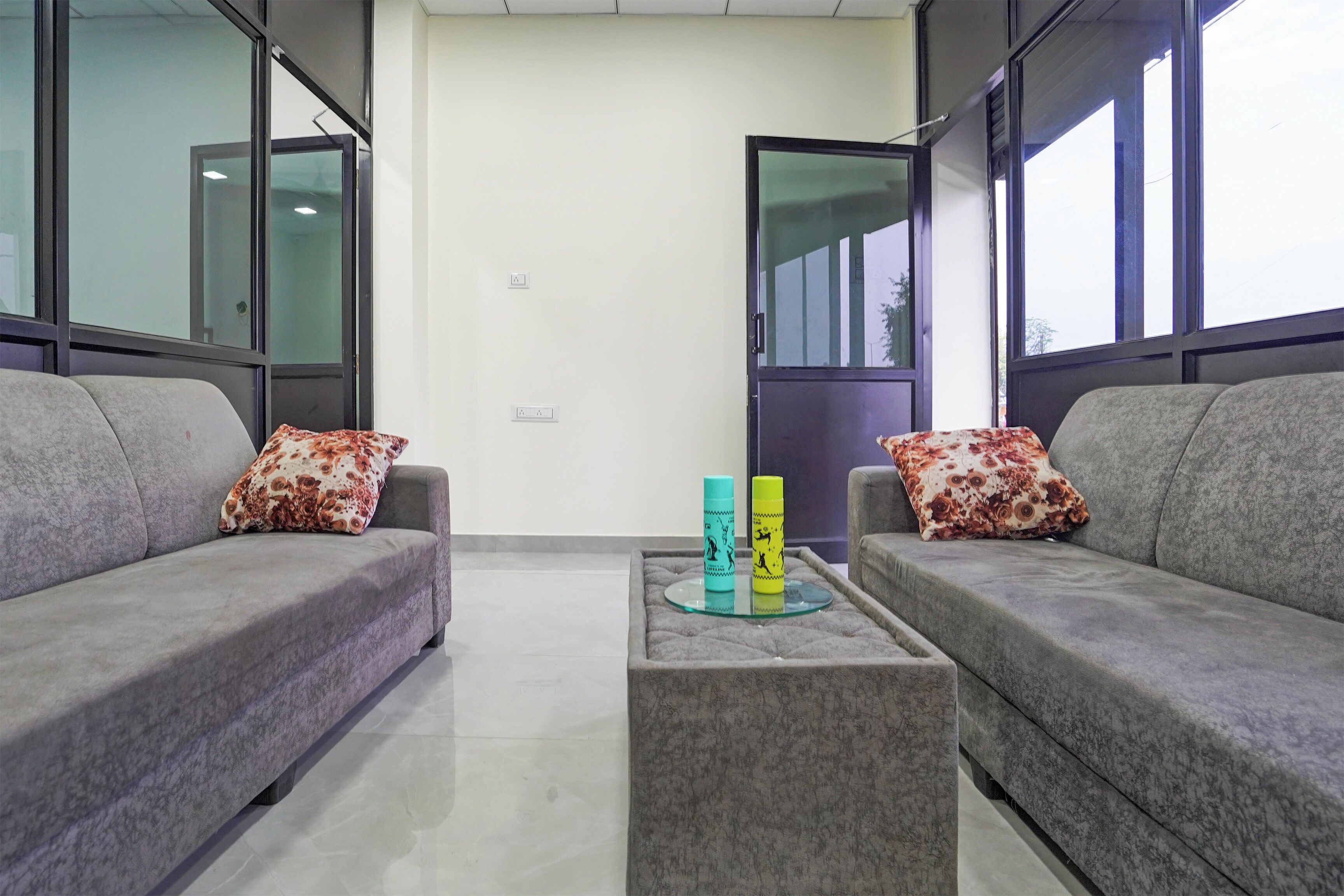 Una sala de estar moderna con dos sofás grises, cada uno con un cojín floral, una mesa de centro de vidrio con dos botellas coloridas y ventanas amplias a ambos lados.
