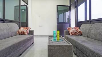 Una sala de estar moderna con dos sofás grises, cada uno con un cojín floral, una mesa de centro de vidrio con dos botellas coloridas y ventanas amplias a ambos lados.