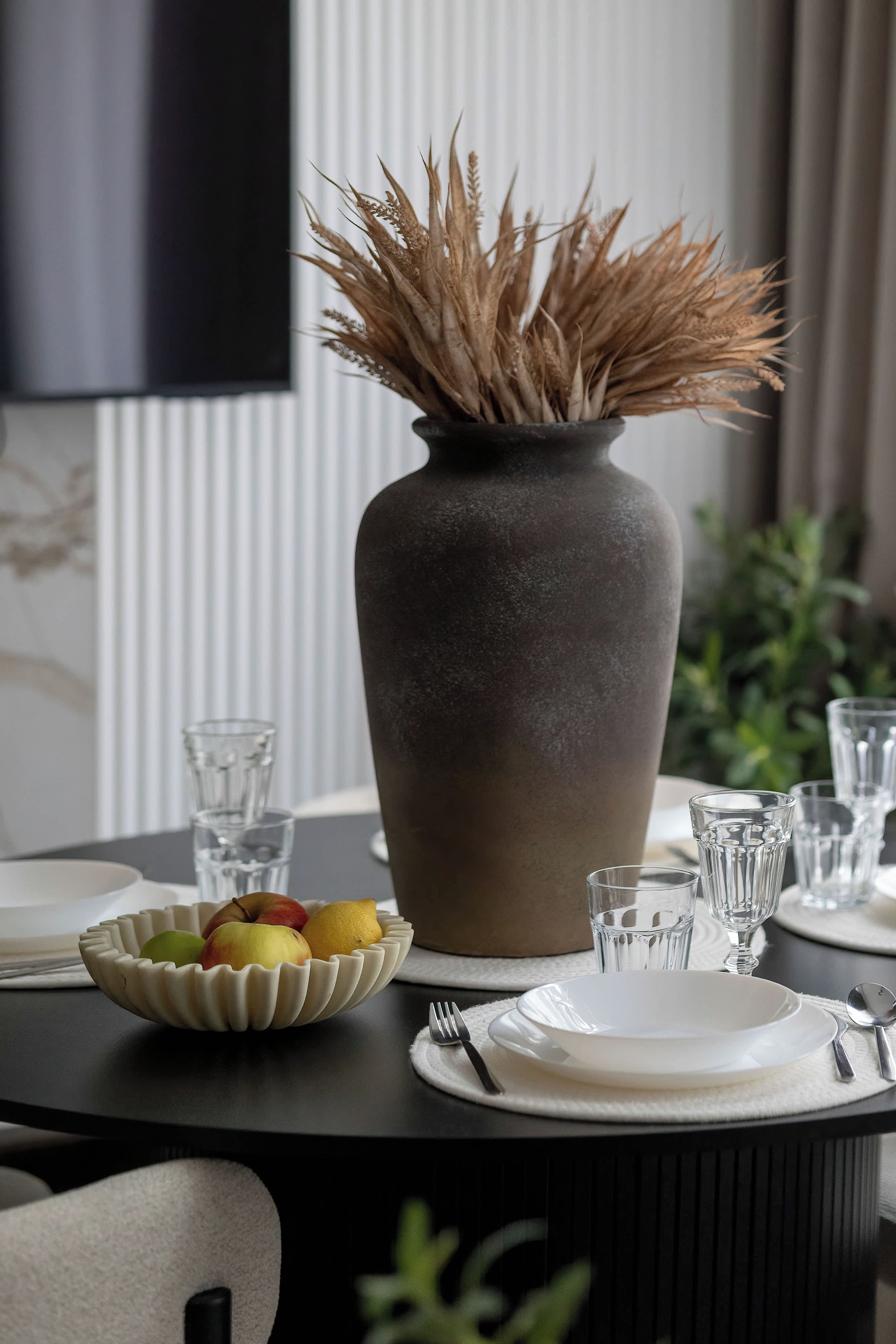 A dining table with a vase of dried plants, a fruit bowl, and clear glasses.