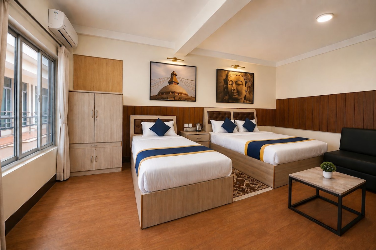 A hotel room with two beds, a desk, a sofa, a window with curtains, and artwork on the walls.