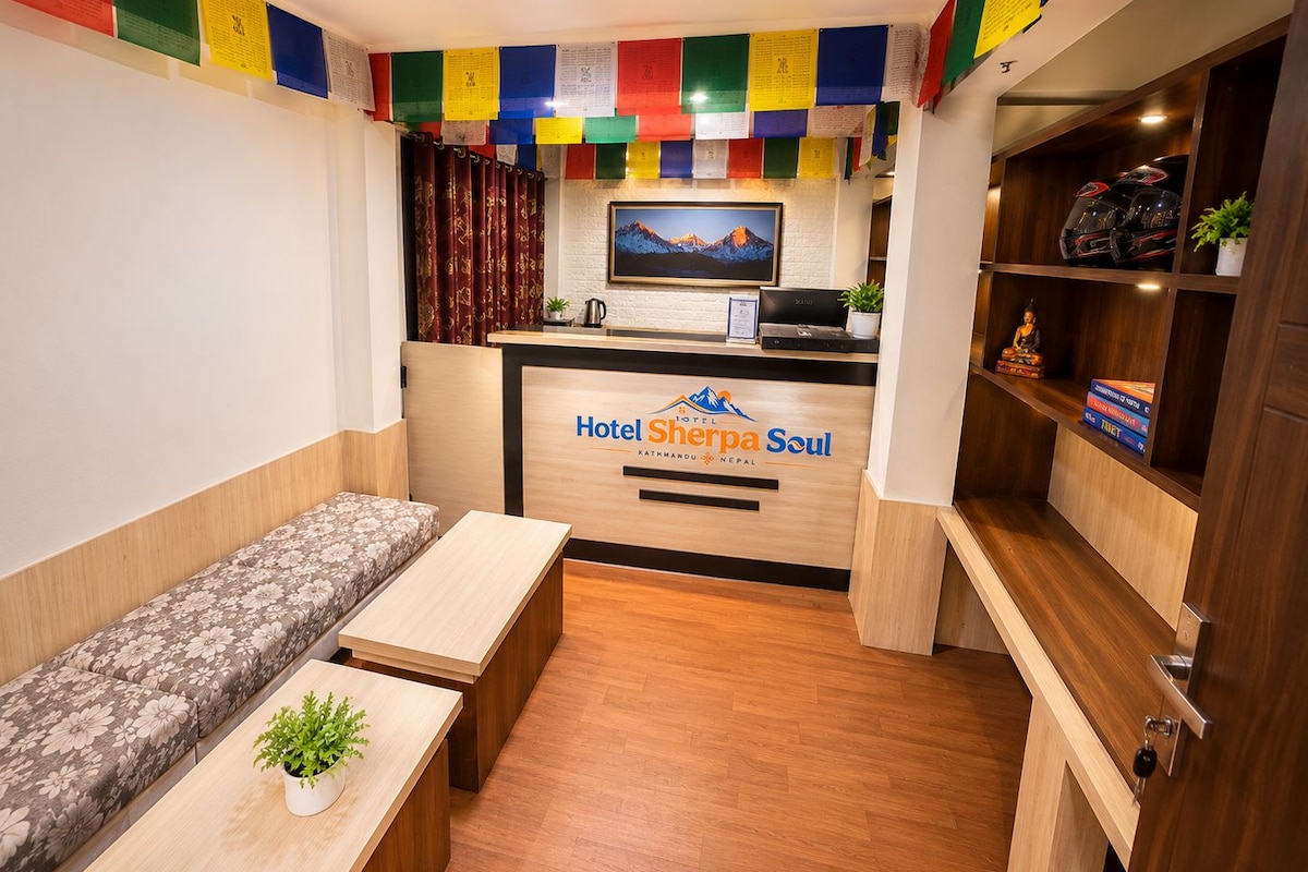 A hotel reception area with a sofa, a desk, a computer, and a shelf with helmets and books.
