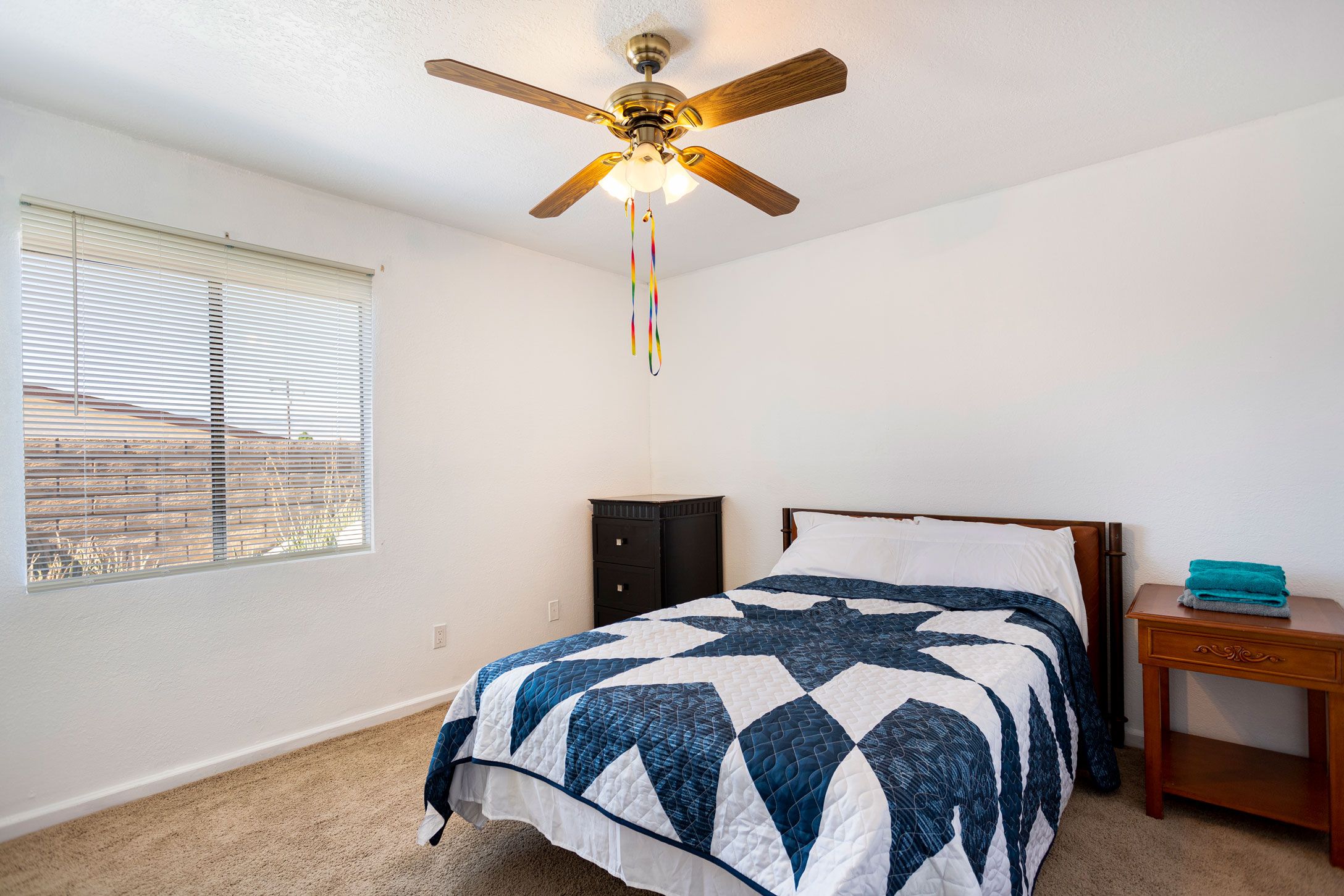 A bedroom with a bed, a ceiling fan, a window with blinds, a nightstand, and a dresser.