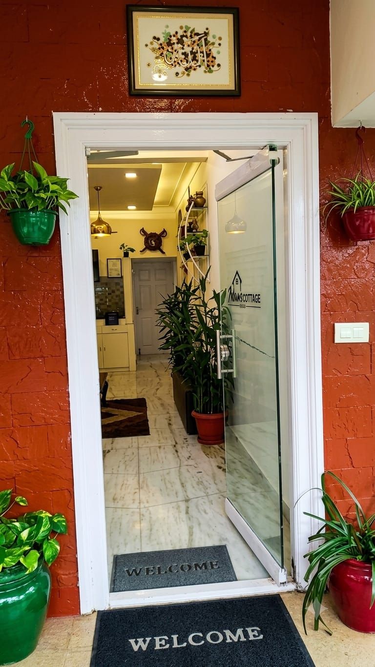 A doorway leading to a kitchen area with a 'Welcome' mat, potted plants, and a glass door with a 'NAN'S COTTAGE' sign.