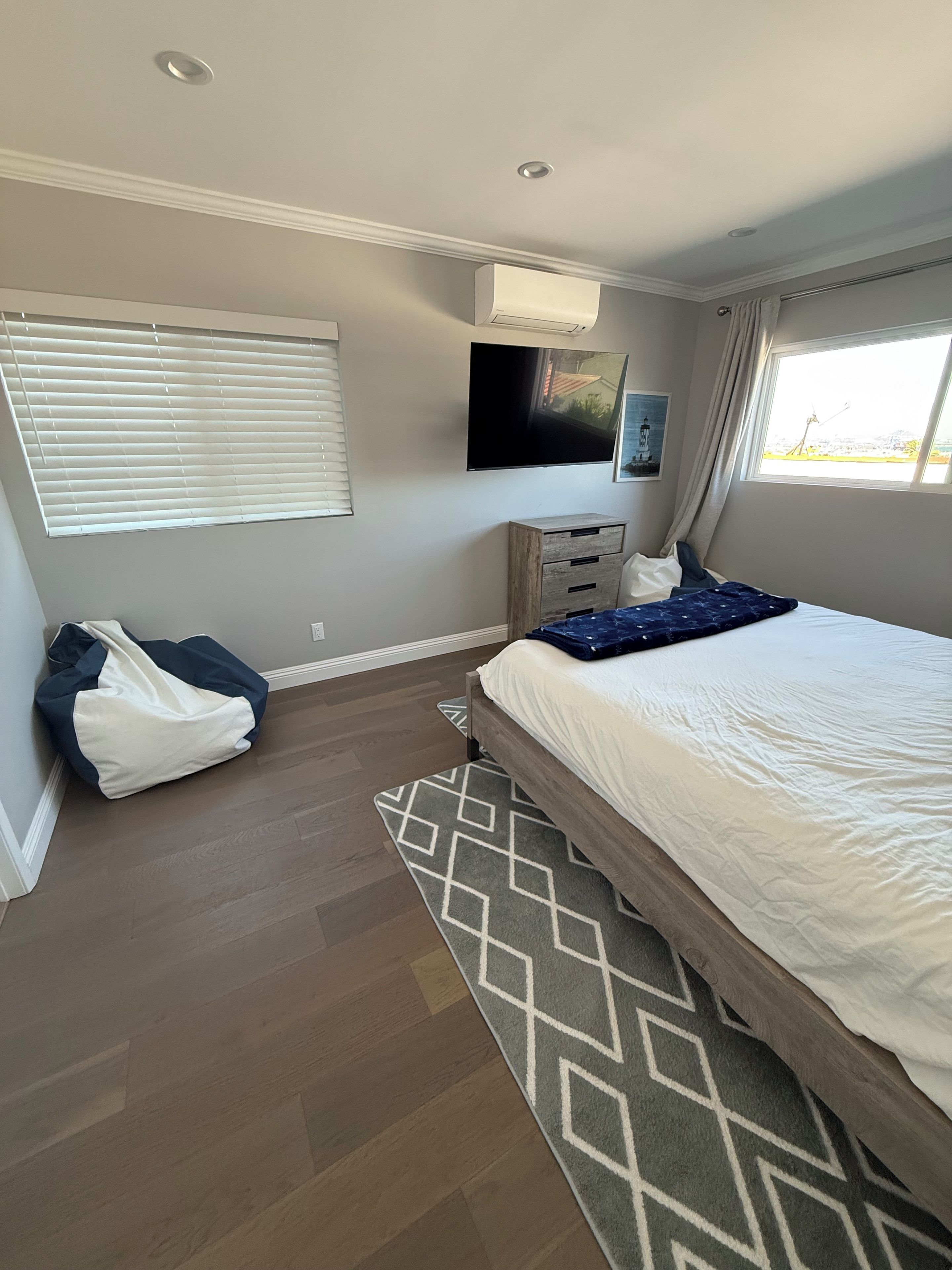 A bedroom with a bed, a TV mounted on the wall, a window with curtains, and a wooden floor.