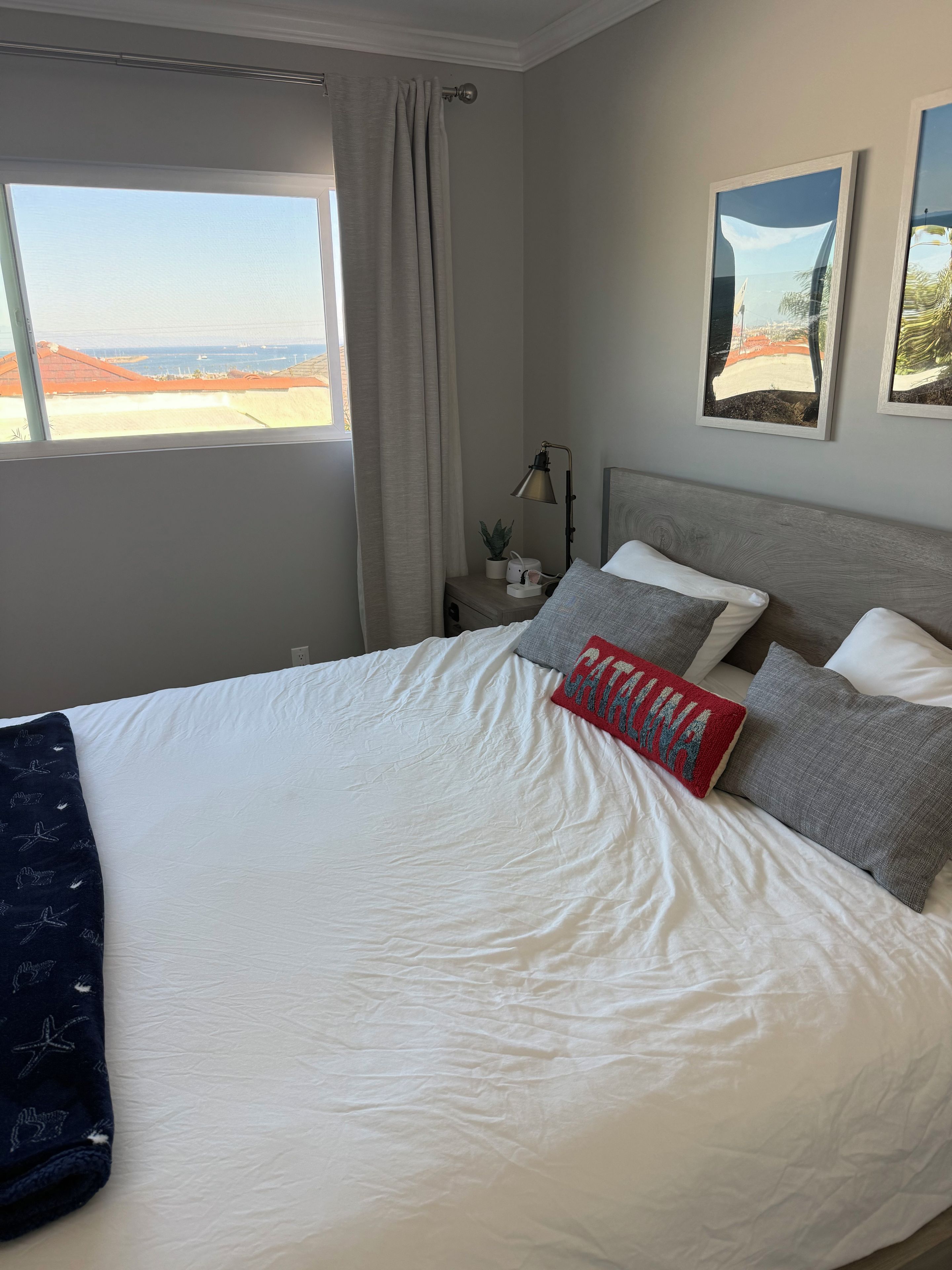 Una cama bien hecha con almohadas y una colcha, una mesita de noche con una lámpara y una planta, y una ventana con vistas al paisaje.