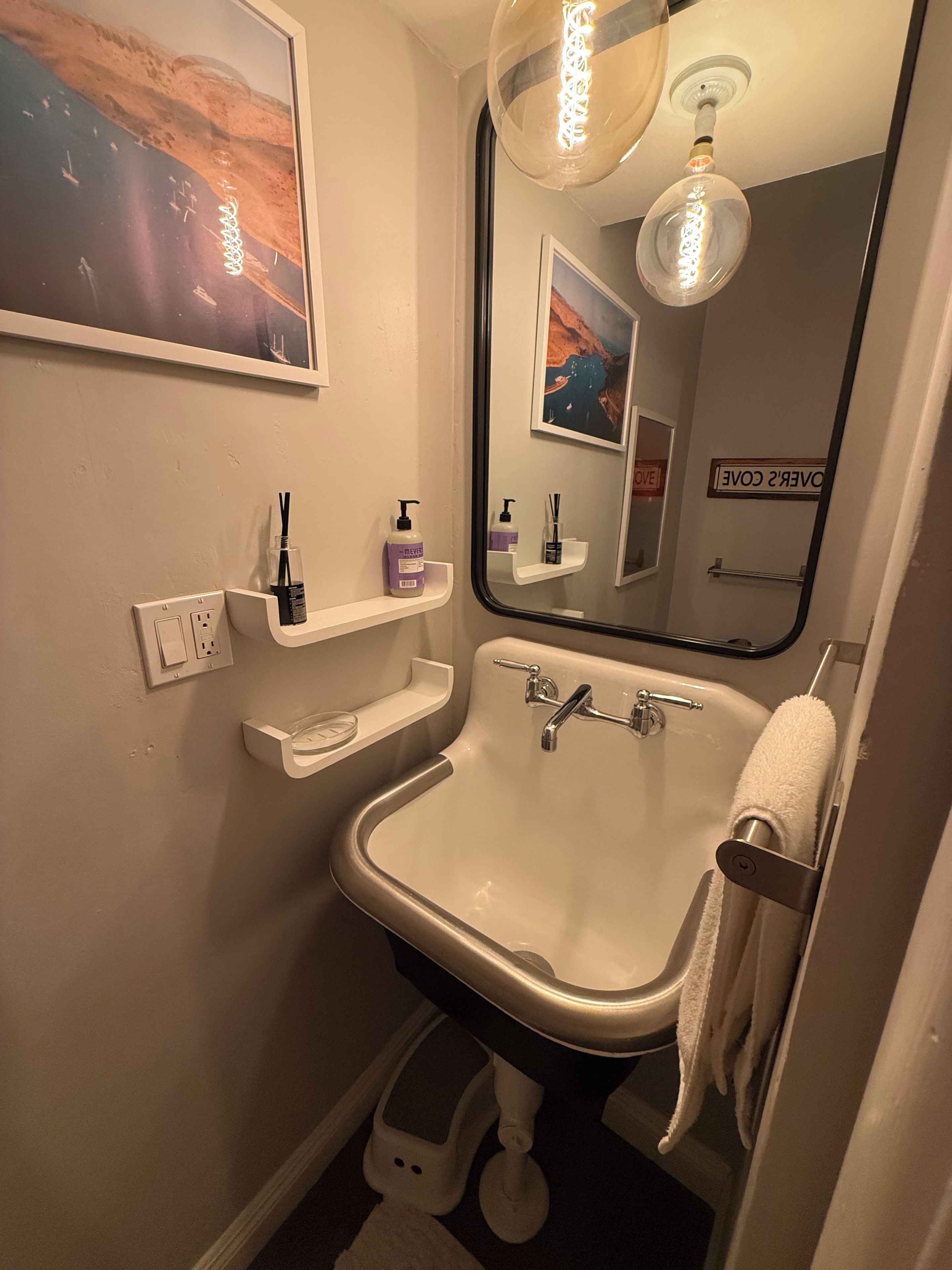 Un bagno con un lavandino bianco, uno specchio, un porta asciugamani e uno scaffale con articoli da toilette.