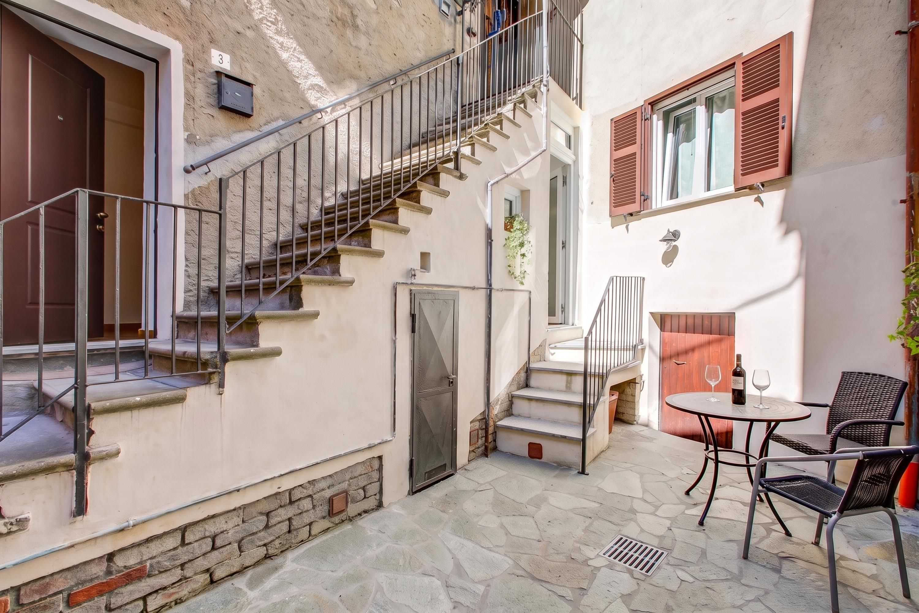Un patio con suelo de piedra, una mesa y sillas, una escalera con barandillas de metal y una puerta con un pequeño ventanal.