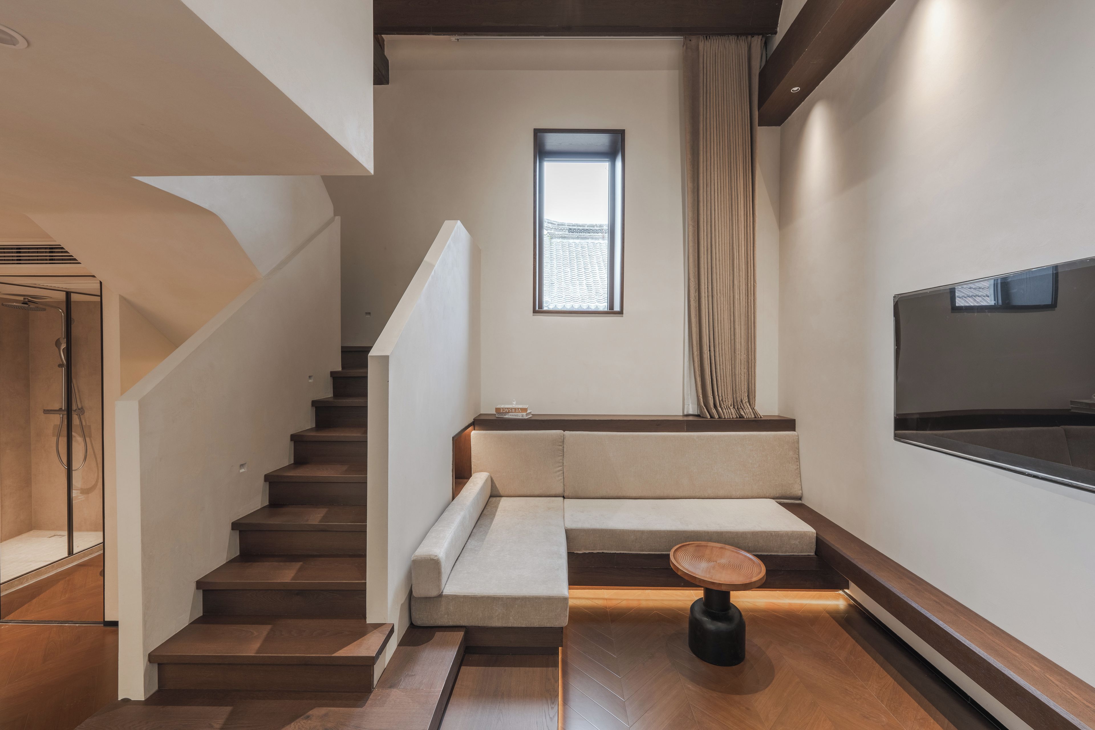 Un escalier menant à un palier où se trouvent un banc rembourré et une petite table, une fenêtre avec des rideaux et un téléviseur à écran plat fixé au mur.