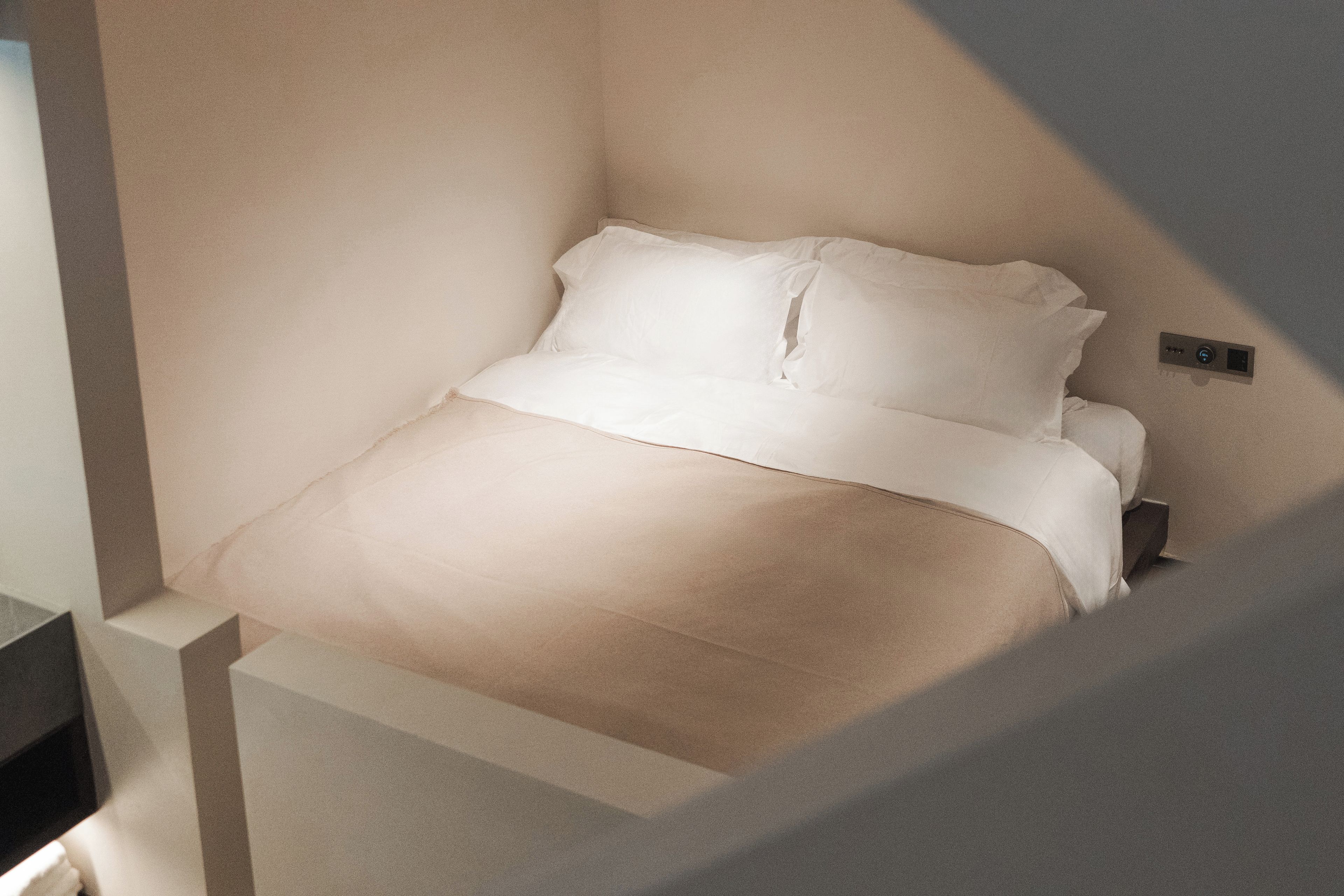 A bed in a corner of a room with a light-colored wall and a small control panel on the wall.