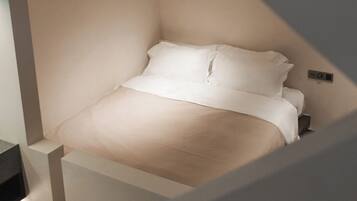 A bed in a corner of a room with a light-colored wall and a small control panel on the wall.