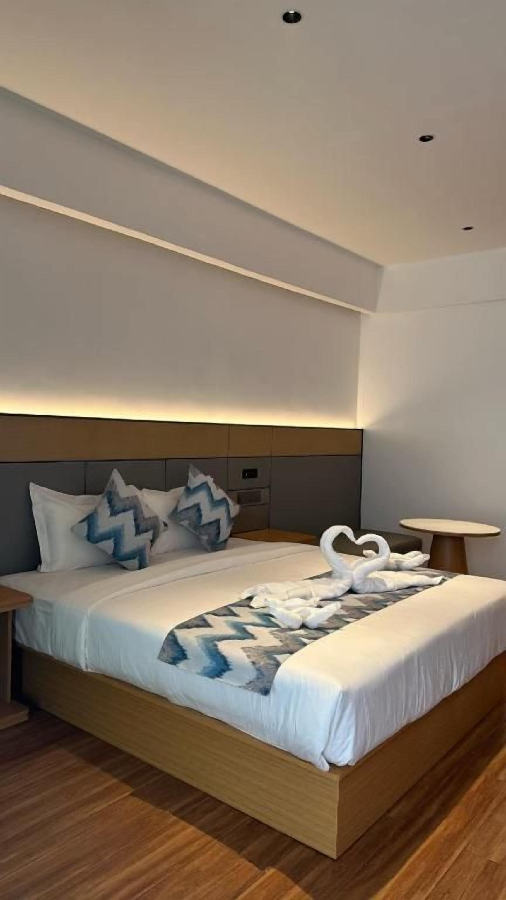 A modern hotel room with a wooden bed frame, a headboard with built-in lighting, and a bedside table with a lamp.