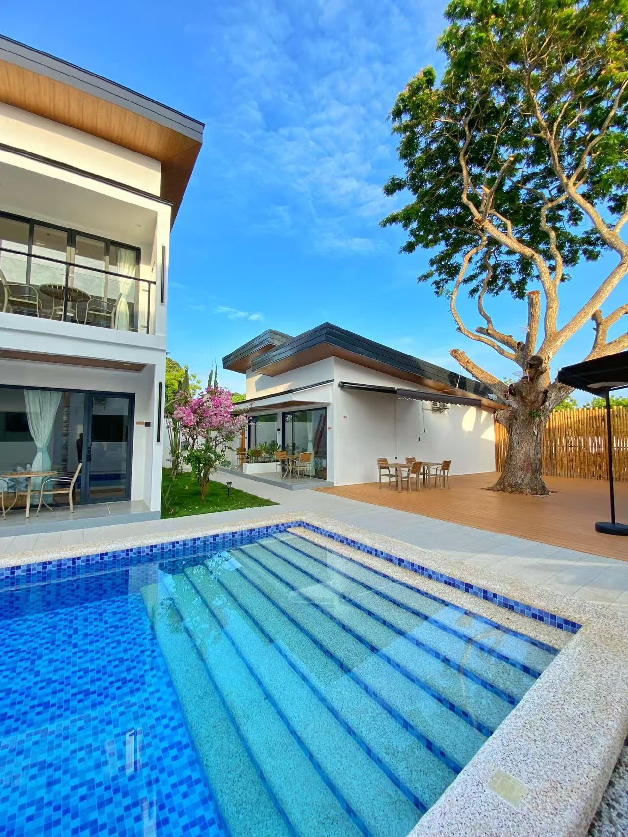 A modern two-story building with a swimming pool, outdoor seating, and a large tree.