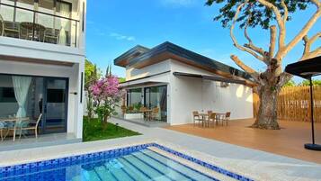 A modern two-story building with a swimming pool, outdoor seating, and a large tree.