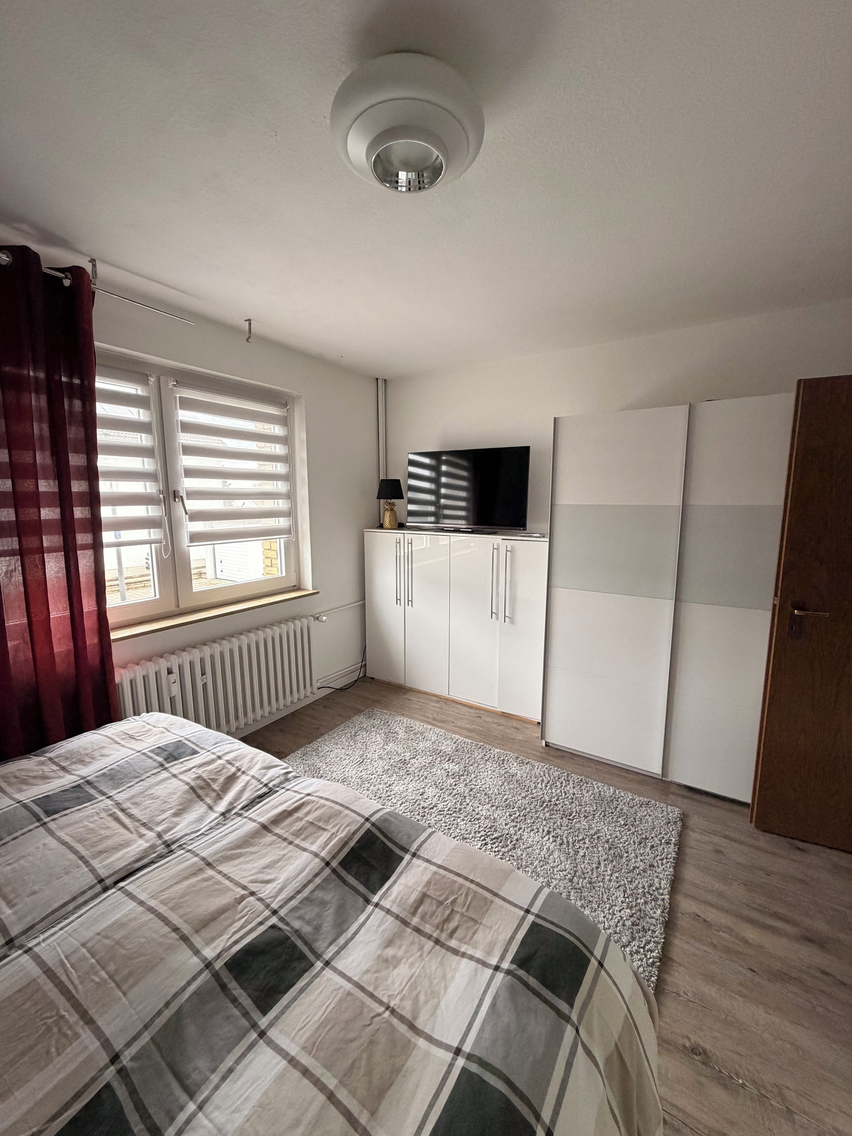 A bedroom with a bed, a radiator, a window with curtains, a wardrobe, and a television on a cabinet.