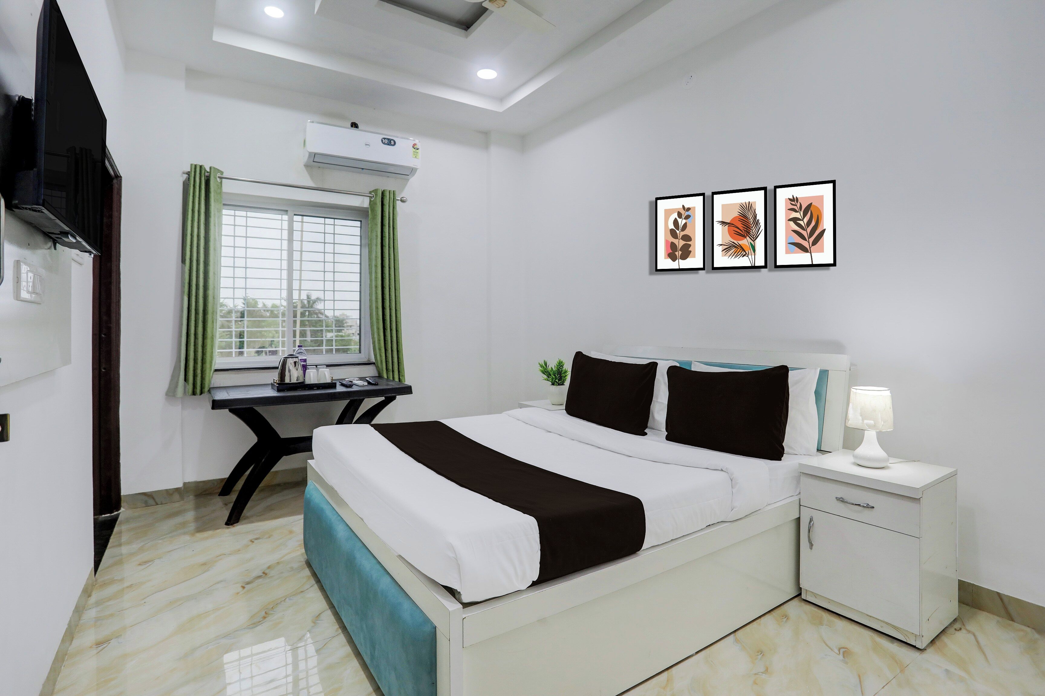 A hotel room with a bed, bedside table, nightstand, and a desk with a chair. There is a window with curtains, an air conditioning unit, and three framed pictures on the wall.