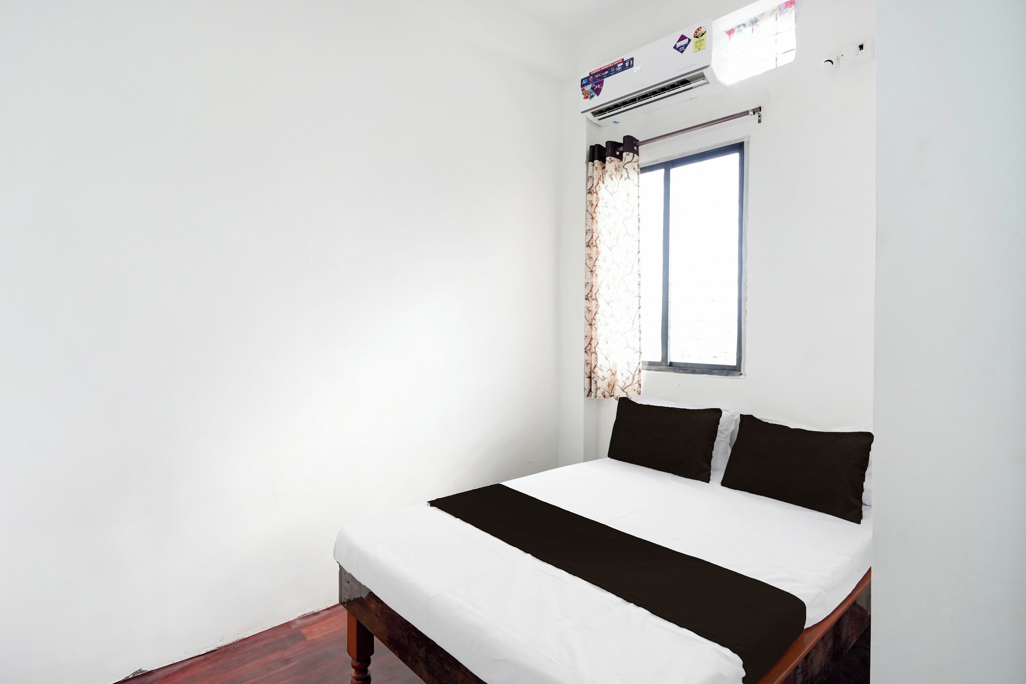 Ein Schlafzimmer mit einem Bett, weißen Wänden, einem Fenster mit Vorhängen und einer Klimaanlage.