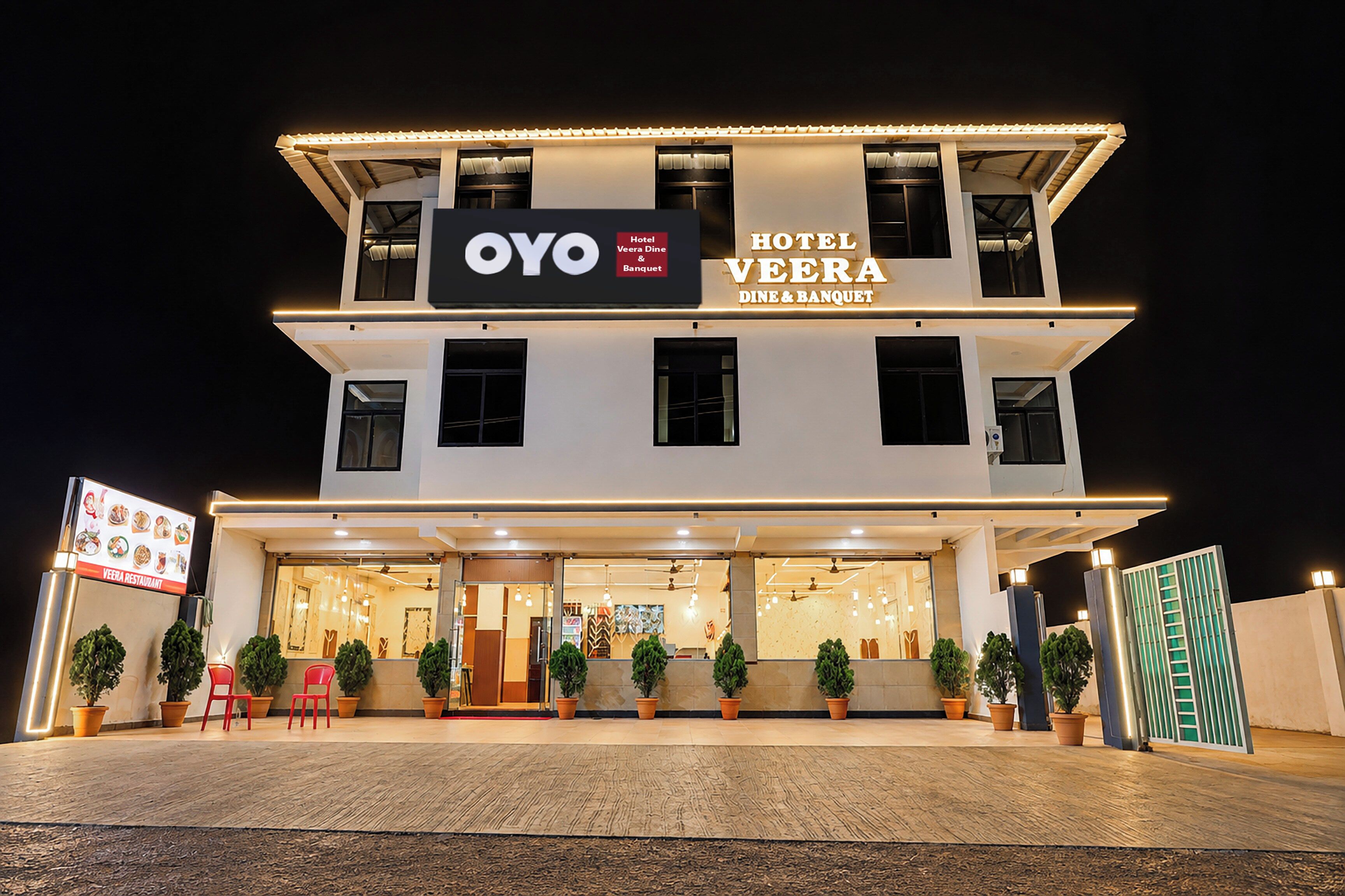 A two-story hotel with a sign for 'OYO Hotel Veera Dine & Banquet' and a visible entrance with potted plants and red chairs outside.