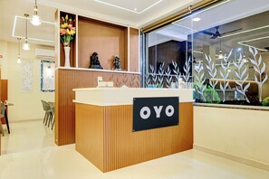 Balcão de recepção com placa indicando 'OYO', em ambiente moderno com painel de madeira e uma grande janela com folhagem decorativa.