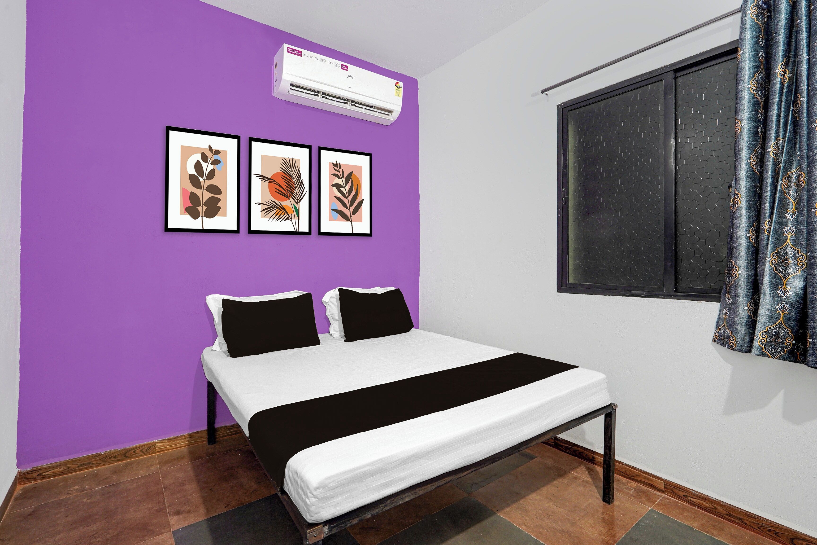 A bedroom with a bed, a window with curtains, an air conditioner, and three framed pictures on the wall.