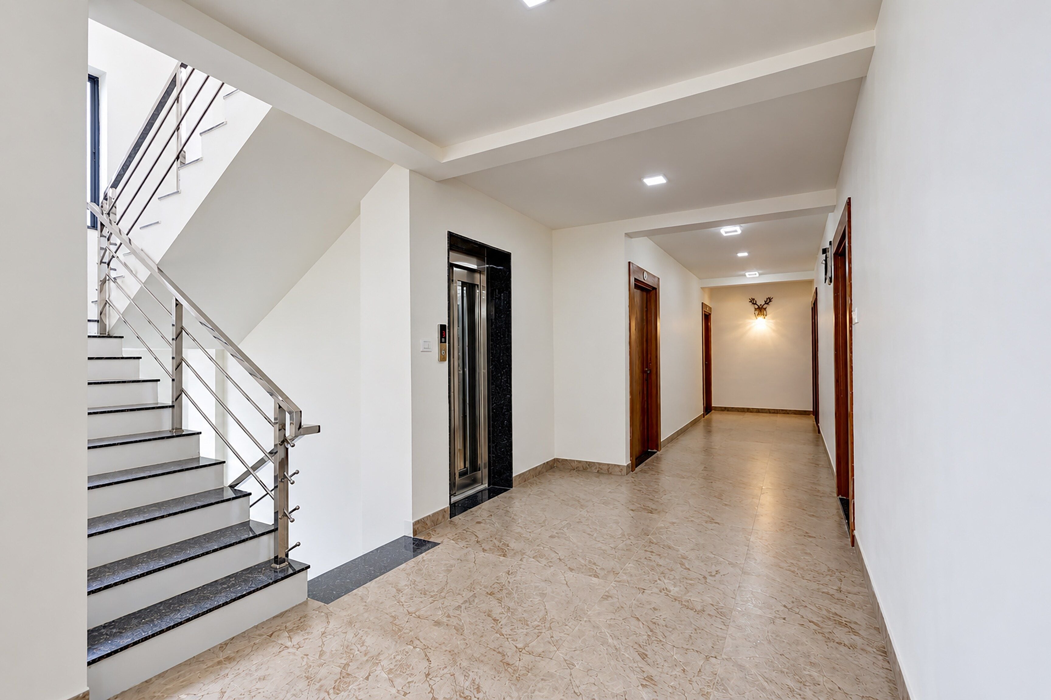 Un interior moderno con una escalera, un ascensor y un pasillo que conduce a una puerta con una lámpara de pared.