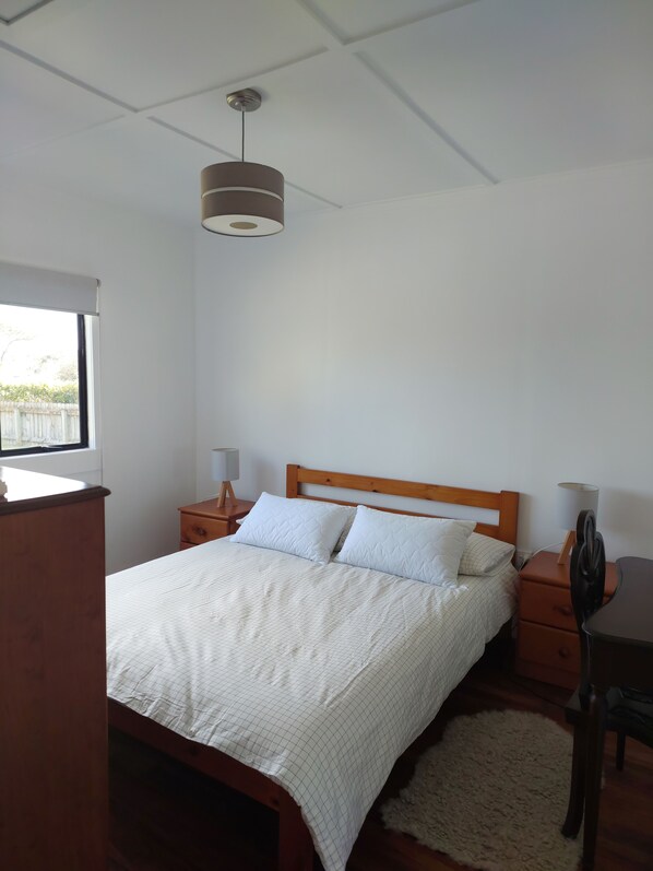 Un dormitorio con cama, cabecera de madera, mesitas de noche, lámpara, escritorio y ventana.