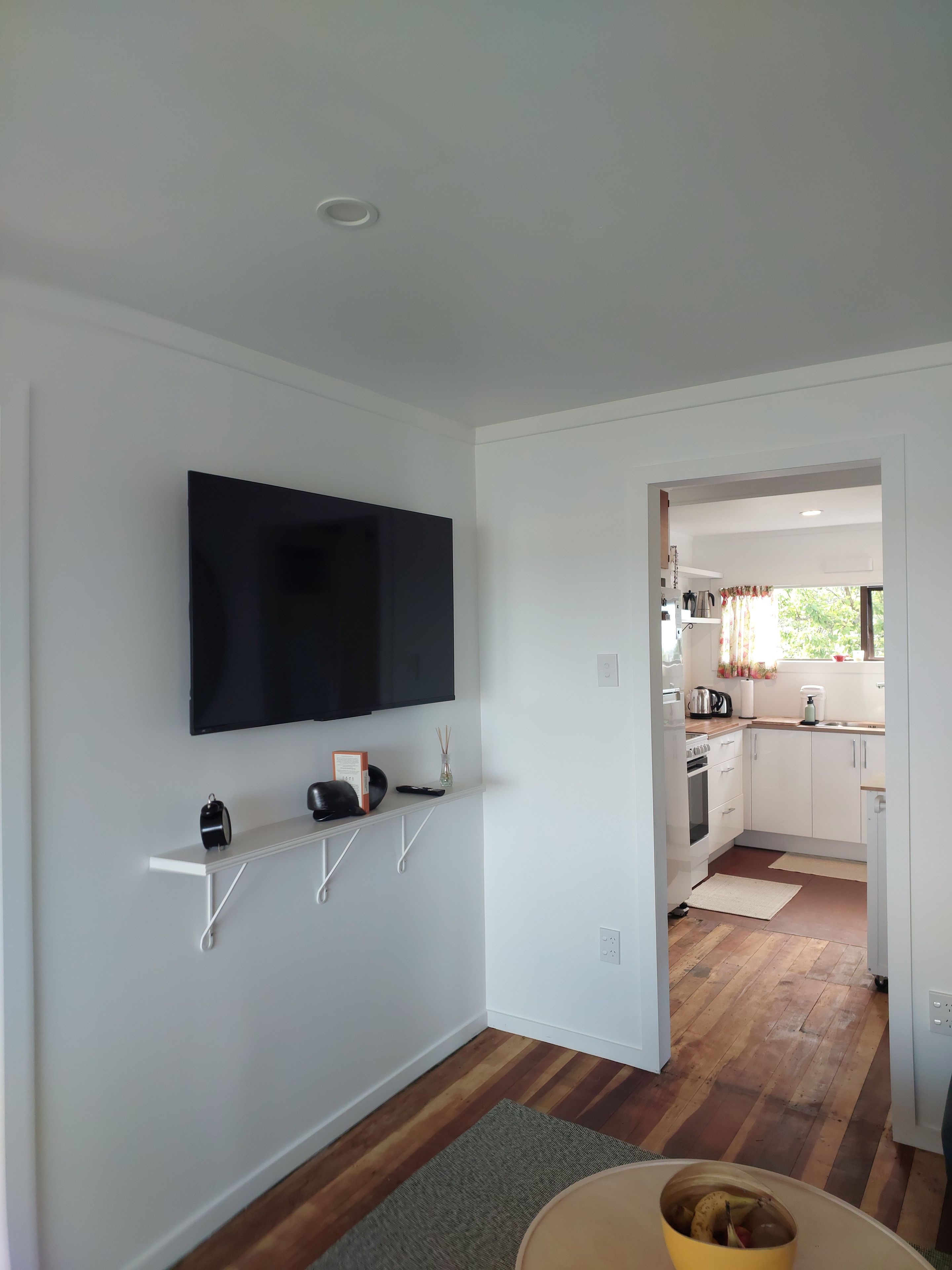 Un salon moderne avec un téléviseur fixé au mur, des étagères blanches, un plancher en bois et une vue sur la cuisine.