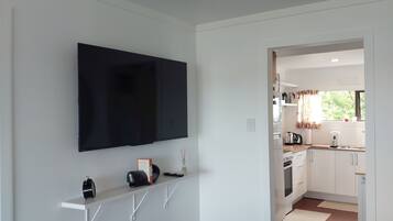 Un salon moderne avec un téléviseur fixé au mur, des étagères blanches, un plancher en bois et une vue sur la cuisine.