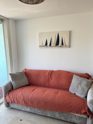 A sofa with a red blanket and two pillows, a painting of sailboats on the wall, and a window with blinds. - New Listing (Canet-en-Roussillon)