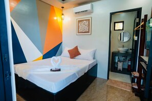 Una camera d'albergo con un letto, un comodino, un condizionatore d'aria a parete, un'opera d'arte incorniciata, uno specchio e un piccolo ripiano con articoli da bagno.