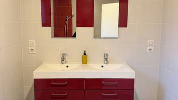 A bathroom with a wooden ceiling, white tiled walls, a double sink vanity with red drawers, and a black trash bin.
