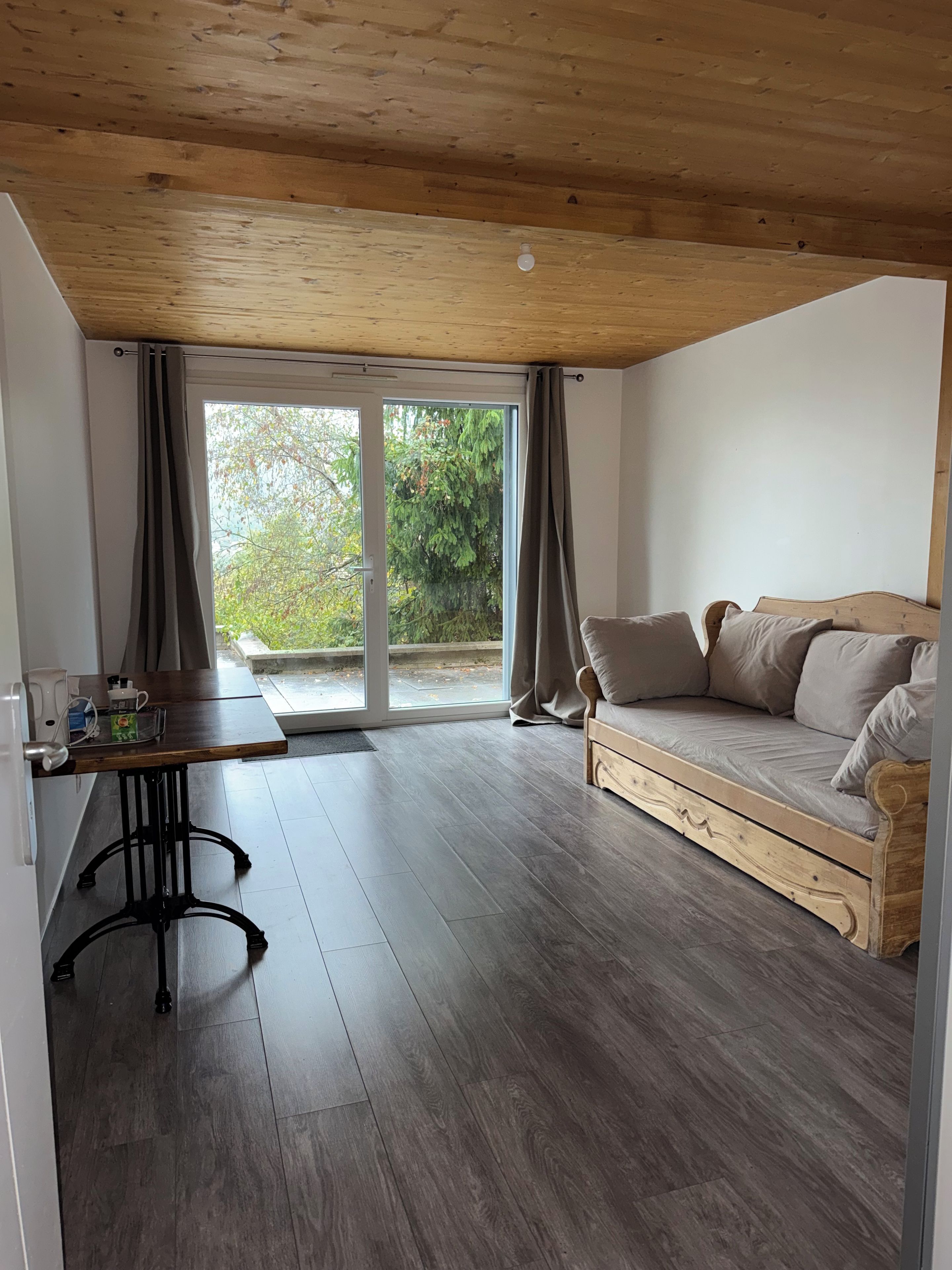 Une pièce avec un plafond en bois, une porte-fenêtre coulissante donnant sur un espace extérieur, un canapé et une petite table avec une bouilloire.