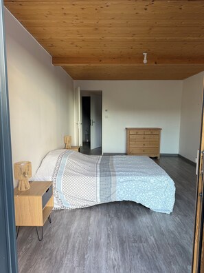 Une chambre à coucher comprenant un lit, une commode en bois, une table de chevet et une porte donnant sur une autre pièce.