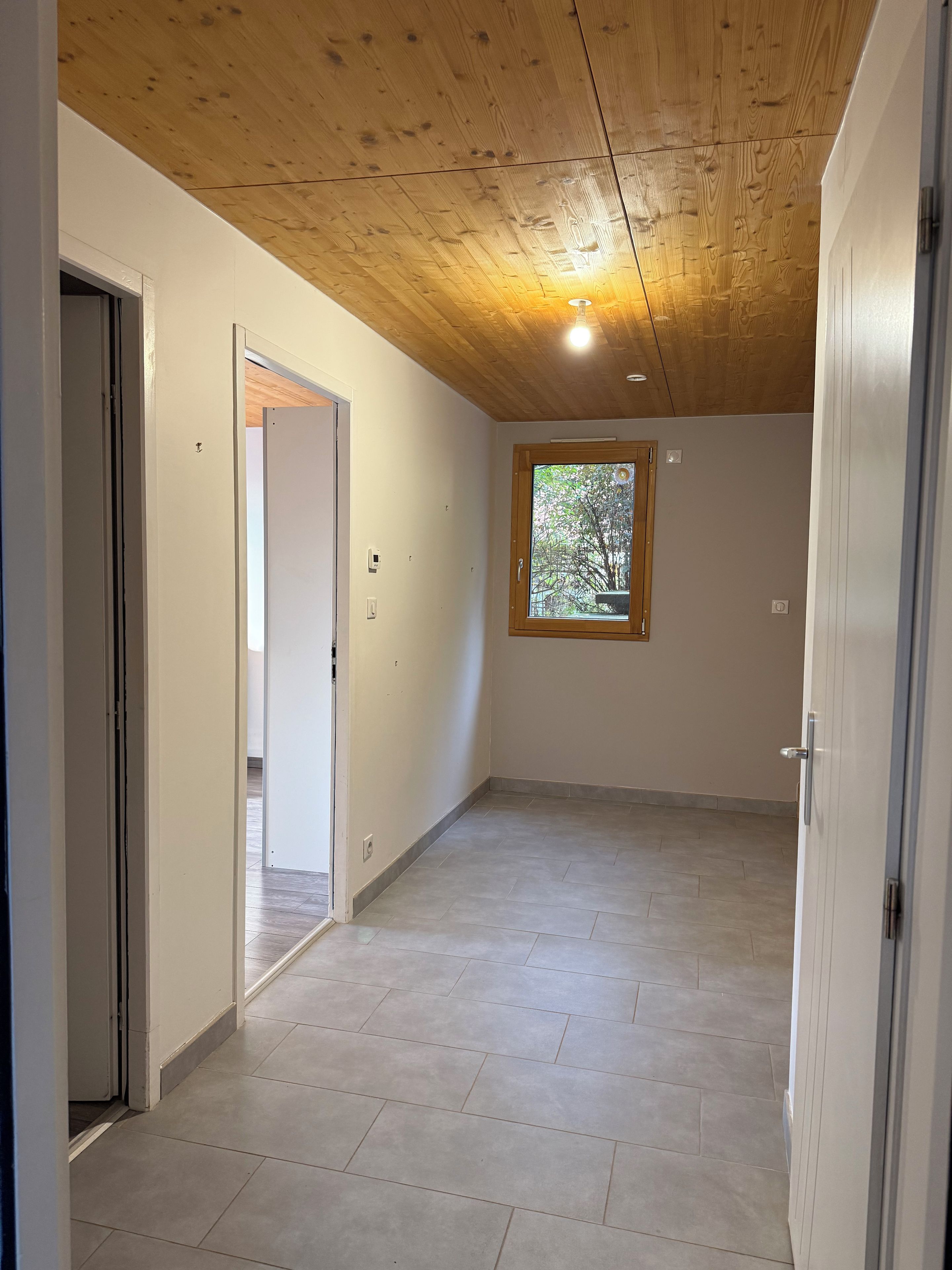 Une pièce avec un plafond en bois, une fenêtre donnant sur les arbres et un plancher carrelé.