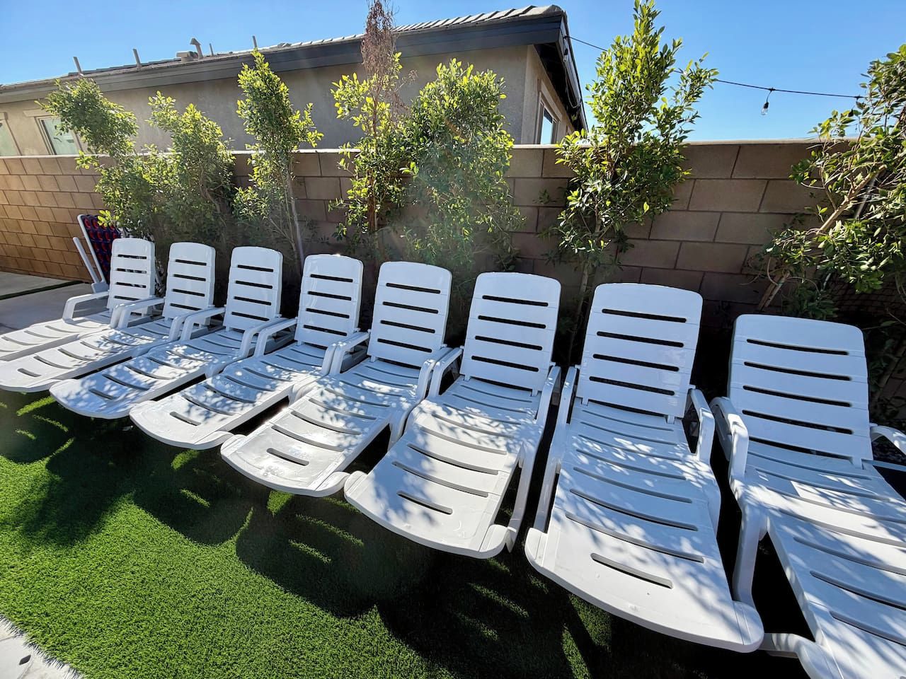 A row of white lounge chairs aligned on a green surface, likely a lawn, with a building in the background.