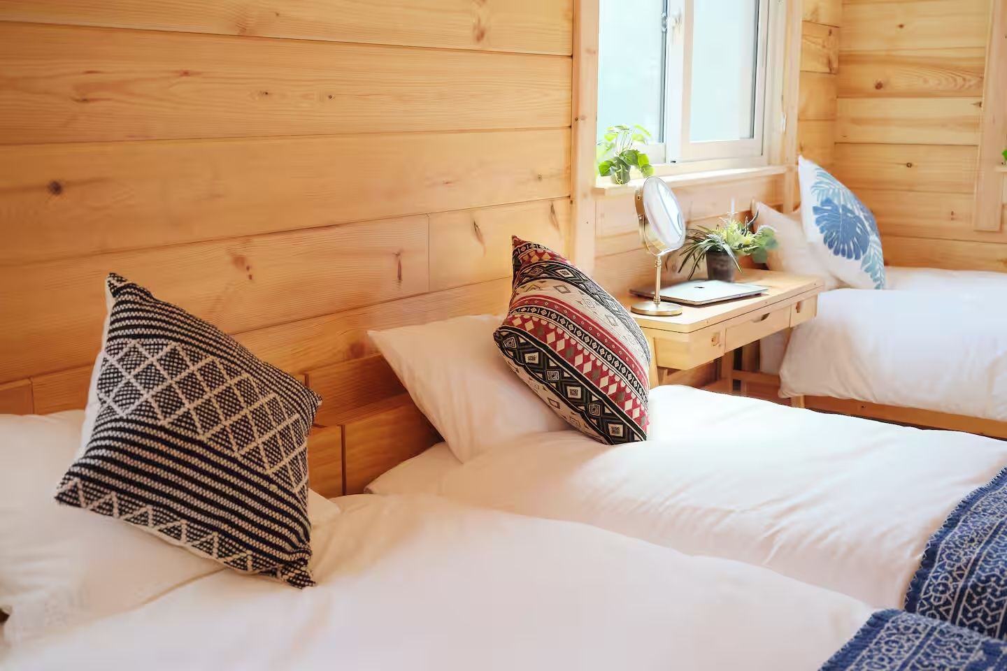 A wooden cabin room with two beds, each with patterned pillows, a small table with a lamp, and a window with a plant.