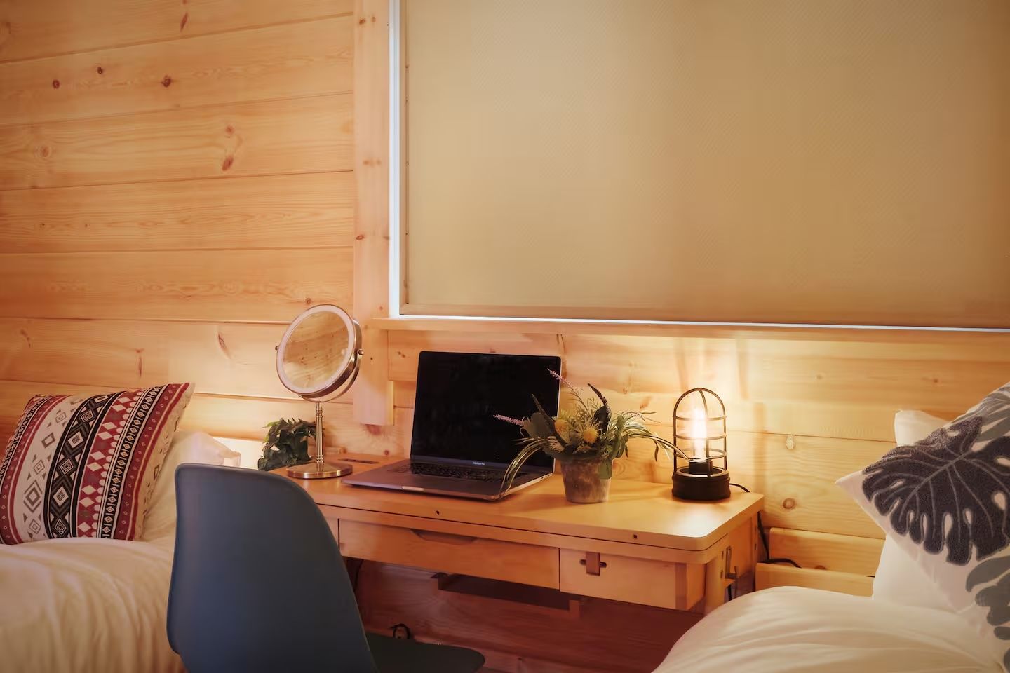 A wooden room with a bed, a desk with a laptop, a mirror, a potted plant, and a decorative light.