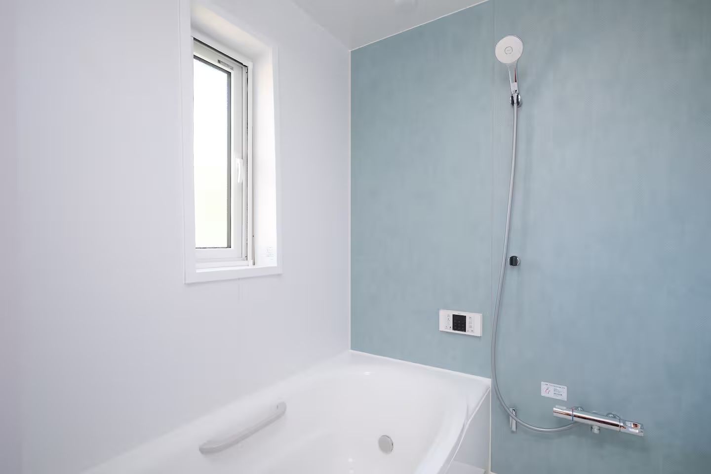 A bathroom with a white bathtub, a shower with a handheld showerhead, a window, and a thermostat on the wall.