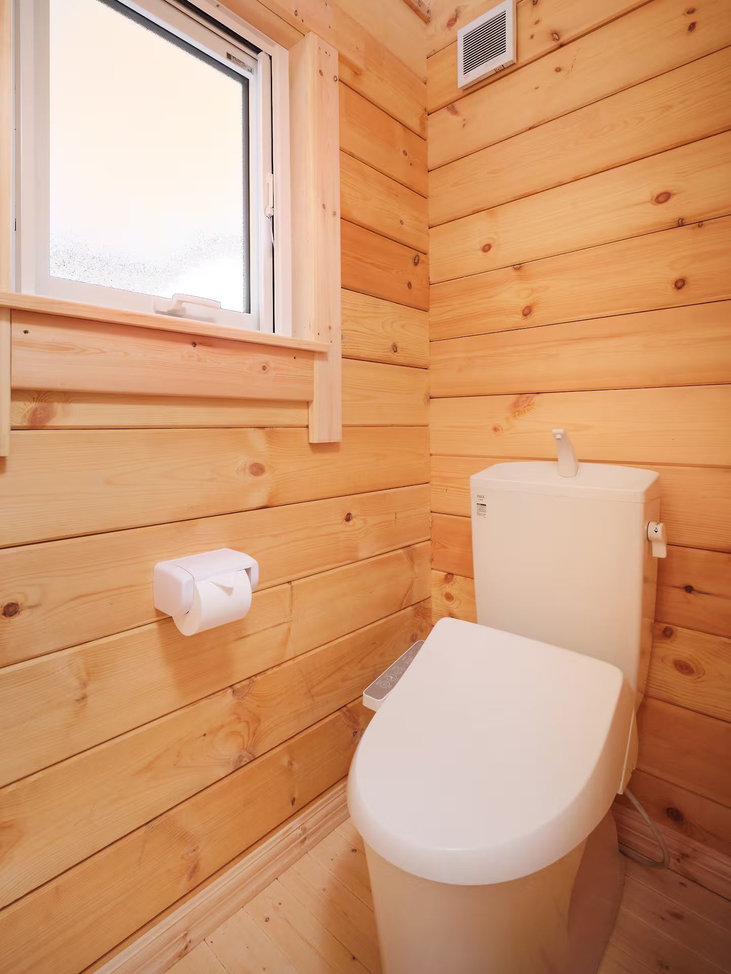 A small, wooden-paneled bathroom with a toilet, a window, and a vent.