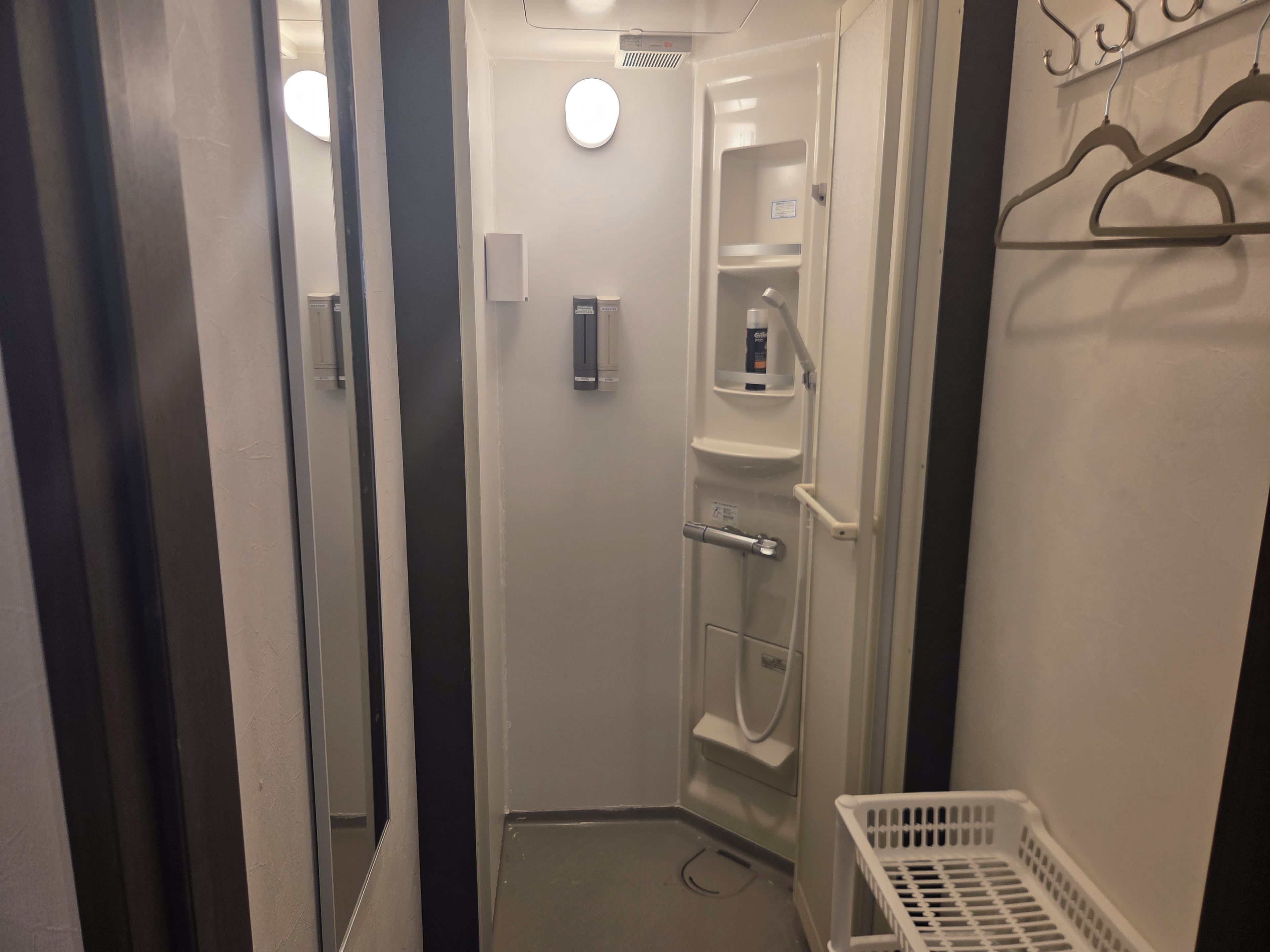A narrow hallway with a door leading to a bathroom. The bathroom has a shower, a toilet, and a small shelf with toiletries. There is a white basket on the floor.