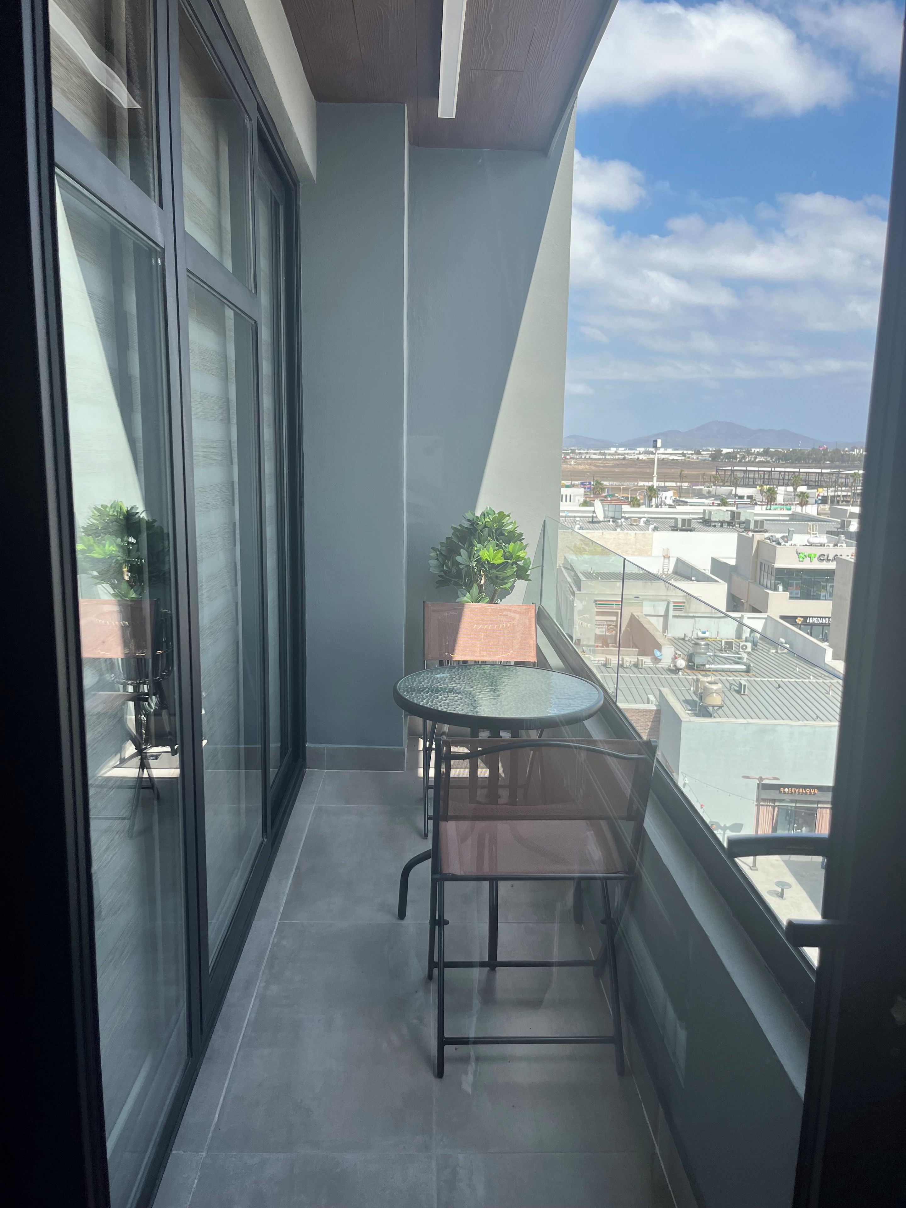 Un balcón con una mesa y sillas de vidrio, una planta en maceta y vistas a la ciudad.