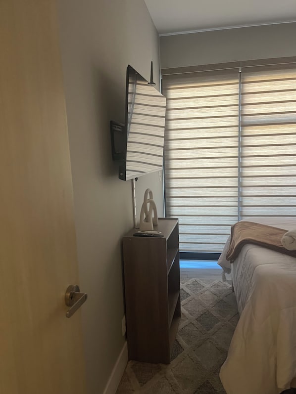 A hotel room with a bed, a bedside table with a lamp, a TV mounted on the wall, and a window with blinds. - New Listing (Tijuana)