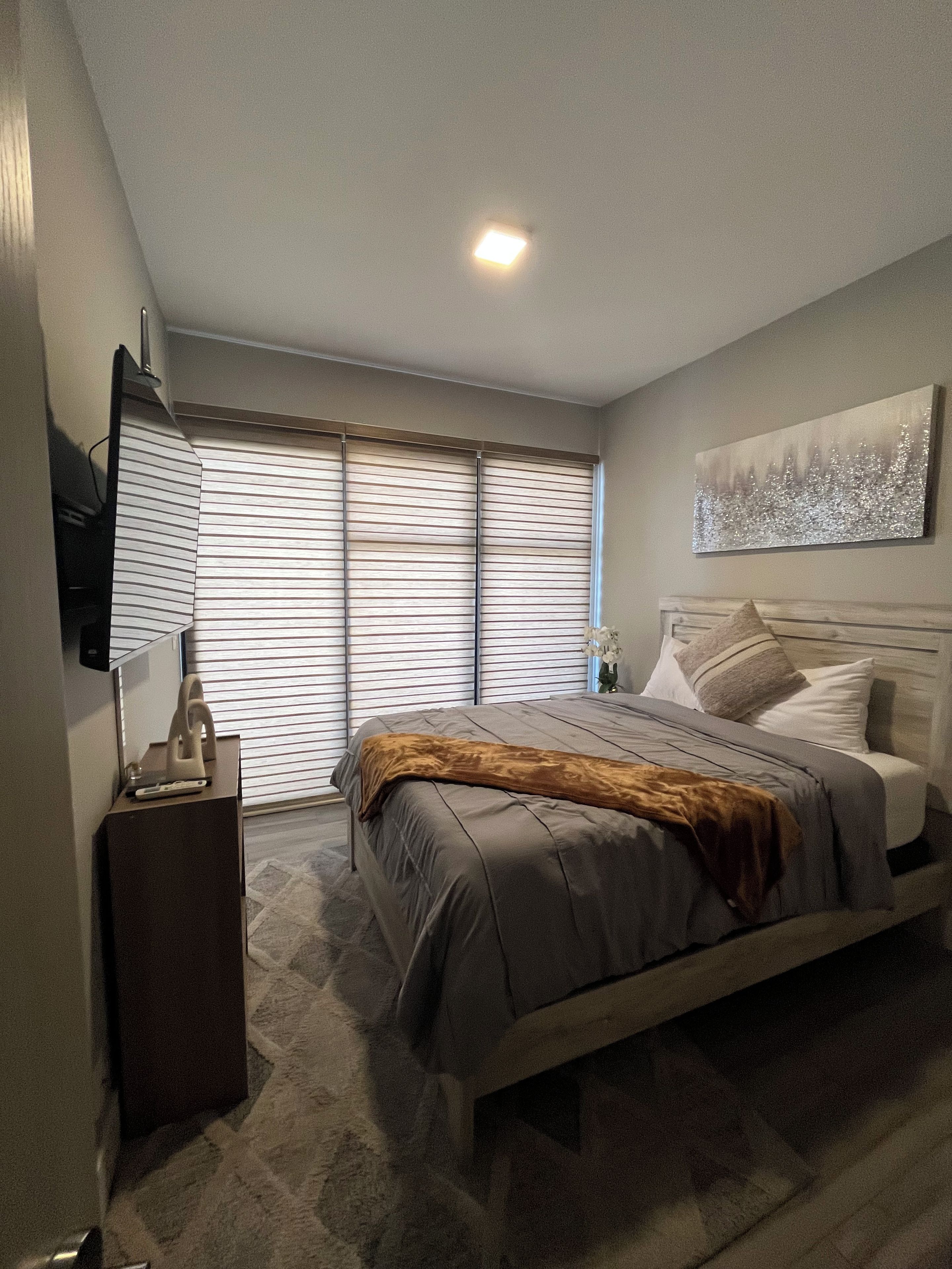 Un dormitorio con cama, mesita de noche, televisor y ventana con persianas.