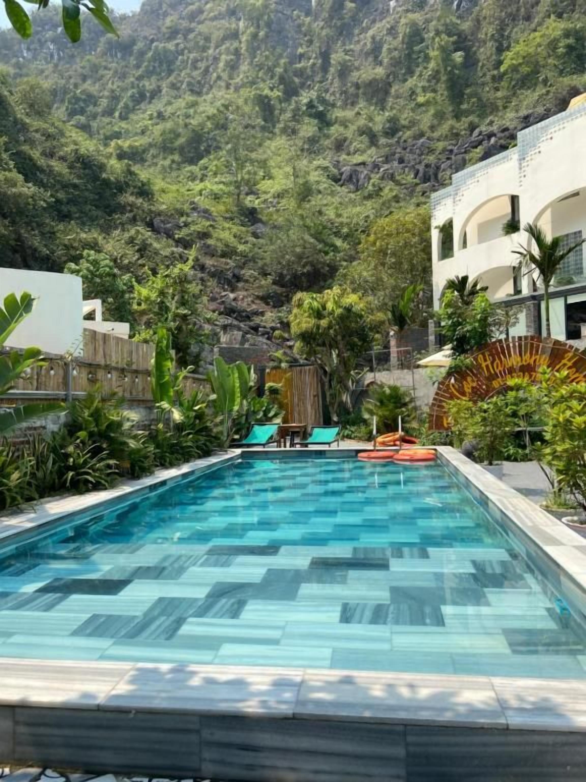 A swimming pool with a view of a mountainous landscape, surrounded by greenery and a building with a white facade.