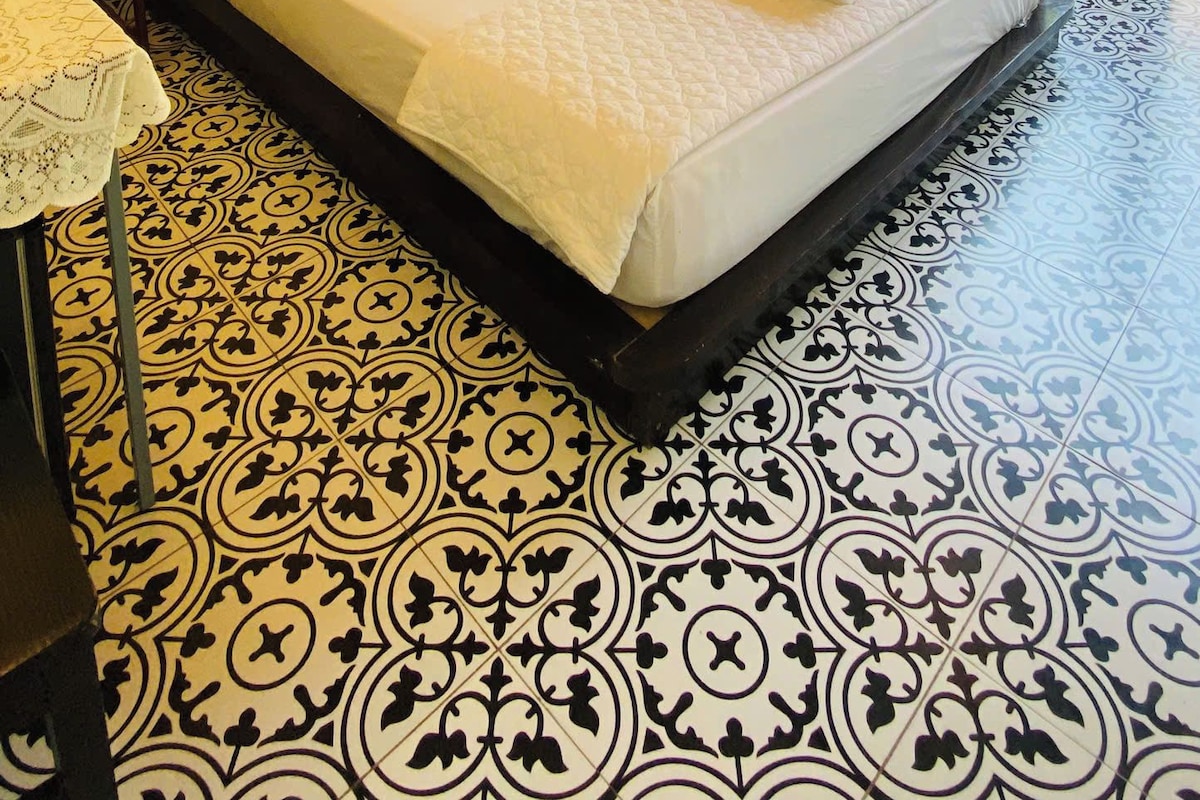 A hotel room with a bed, a nightstand, a lamp, a window with a view of greenery, and a patterned floor.